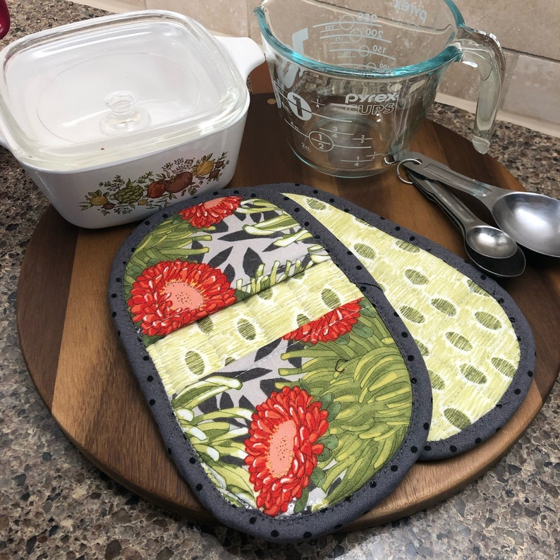 May include: Two gray and white pot holders with a floral pattern. The pot holders have a green, white, and red floral design with a gray border. The pot holders are on a wooden surface.
