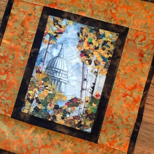 May include: A framed quilted artwork featuring a building with a dome and trees with fall foliage. The background is a light blue watercolor wash. The frame is black with a distressed brown inner border. The outer border is a bright orange and yellow fabric with a mottled pattern.