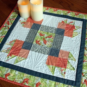 May include: A small, square table runner with a patchwork design featuring red, orange, green, and gray fabrics. The runner has a dark blue border and a quilted pattern.