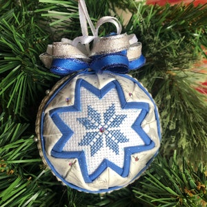 May include: A white and blue quilted Christmas ornament with a snowflake design and silver and blue ribbon.