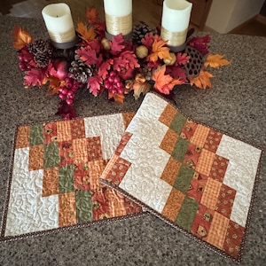 May include: Two rectangular quilted placemats featuring a patchwork design of cream, olive green, and orange fabric squares with a swirling stitch pattern, finished with a dark brown patterned border, displayed with a fall-themed centerpiece.