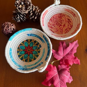 May include: Two round coiled rope bowls. One bowl features a teal, yellow, and red mandala pattern at the center. The other bowl has a coral and orange leaf-like print interior. Both bowls are constructed from off-white cotton rope.