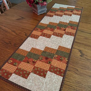 May include: A rectangular table runner with a patchwork design in shades of orange, green, and cream. The runner features a repeating pattern of squares and rectangles, with a patterned border. The runner is approximately 48 inches long.