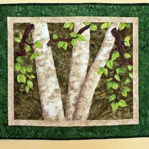 May include: A framed quilted wall art piece depicting three birch trees with green leaves. The trees are white with brown branches and are set against a green background.