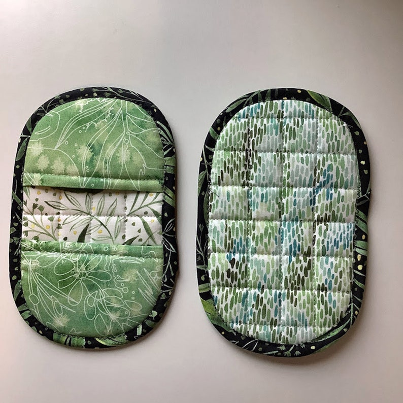 May include: Two green and black quilted pot holders with different patterns. One pot holder has a leafy pattern and the other has a green and blue speckled pattern.
