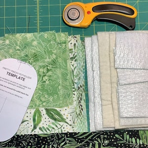 May include: A green and white fabric with a floral pattern, a template for a potholder, a rotary cutter, and a stack of white and silver fabric squares.