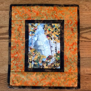 May include: A framed quilted wall hanging with a picture of a white building with a dome roof surrounded by trees with fall foliage. The background is a light blue watercolor wash. The frame is black with a brown outer border. The background of the wall hanging is a bright orange and yellow batik fabric.