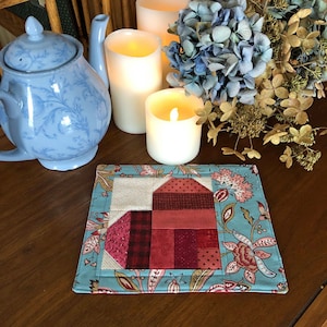 May include: A blue and white floral patterned tea cozy with a red heart design in the center. The cozy is sitting on a wooden table with candles and flowers.