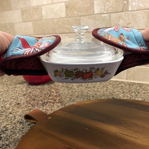 May include: A white casserole dish with a floral design and a glass lid is being held by two red and blue oven mitts. The mitts have a floral pattern and a red polka dot trim.