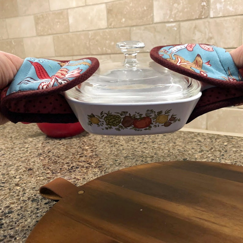 May include: A white casserole dish with a floral design and a glass lid is being held by two red and blue oven mitts. The mitts have a floral pattern and a red polka dot trim.
