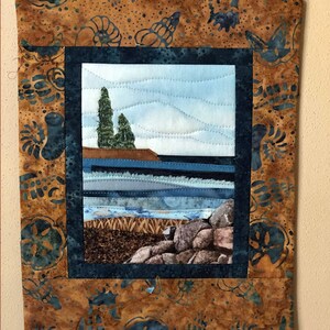 May include: A quilted wall hanging with a brown background and a blue frame. The frame contains a picture of a seascape with two trees on a rocky shore.
