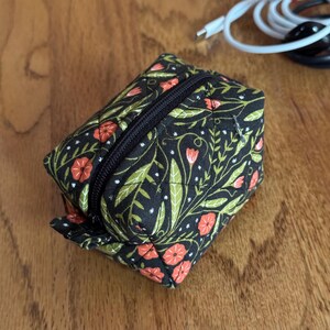 May include: A small, square, zippered pouch with a black background and a floral pattern of green leaves and red flowers. The pouch is quilted and has a black zipper.