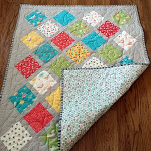May include: A gray and white baby quilt with a diamond pattern. The quilt is made of various colorful fabrics with floral and geometric designs. The quilt is folded in half, revealing a light blue and white patterned backing.