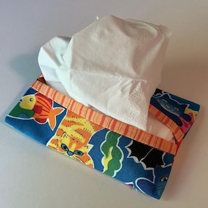 May include: A rectangular tissue holder with a blue fabric featuring orange cats and fish. The top edge has orange and white stripes. White tissues are pulled from the opening.