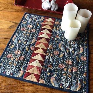 May include: A blue and cream quilted table runner with a geometric pattern of red, cream, and brown triangles. The runner is decorated with a repeating pattern of birds and vines. Three white candles are placed on the runner.