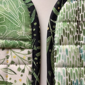 May include: A pair of green and black quilted pot holders with a floral pattern. The pot holders have a black binding around the edges.