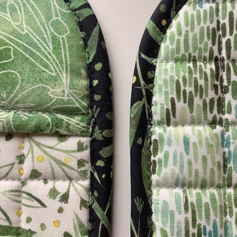 May include: A pair of green and black quilted pot holders with a floral pattern. The pot holders have a black binding around the edges.