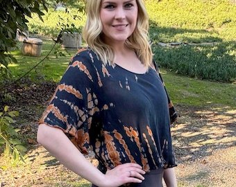 May include: A woman wearing a black and orange tie-dye top with a black cropped top and black leggings. She is wearing gray ankle boots and standing on a gravel surface.