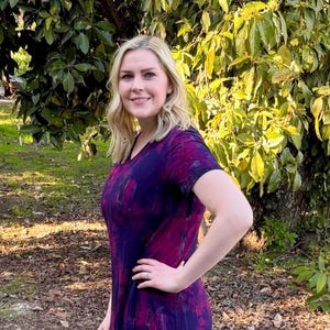 May include: A woman wearing a purple and black tie-dye shirt with black leggings and gray ankle boots. She is standing in a gravel driveway with green grass and trees in the background.