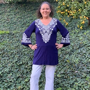 May include: A woman wearing a navy blue tunic top with white embroidered details and white pants. The top has a V-neck and bell sleeves.