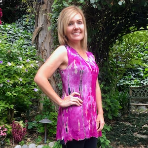 May include: A woman wearing a pink and black tie-dye sleeveless tunic top with a V-neckline. She is wearing black leggings and black heeled sandals. The woman is standing in a garden setting.