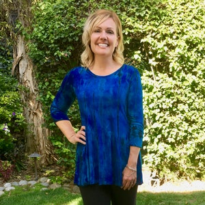 May include: A woman wearing a blue tie-dye top with a long sleeve and black leggings. She is standing in a grassy area and smiling at the camera.