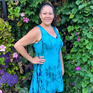 May include: A woman wearing a sleeveless blue tie-dye dress with a V-neckline. She is standing in front of a green leafy background.