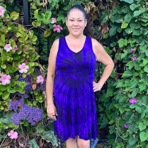 May include: A woman wearing a purple and black tie-dye sleeveless dress with a v-neck. She is standing in front of a green leafy background with pink flowers.