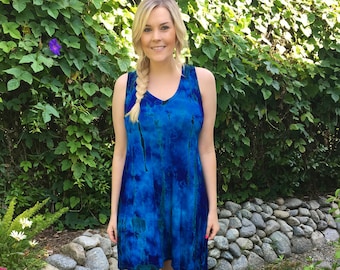 May include: A woman wearing a blue tie-dye sleeveless dress and brown cowboy boots. She is standing in front of a stone wall and green plants.