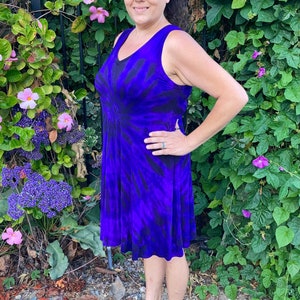 May include: A woman wearing a purple and black tie-dye sleeveless dress with a swirling pattern. She is standing in front of a green leafy background.