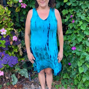 May include: A woman wearing a blue and green tie-dye sleeveless dress with a V-neck. She is standing in front of a green leafy background.