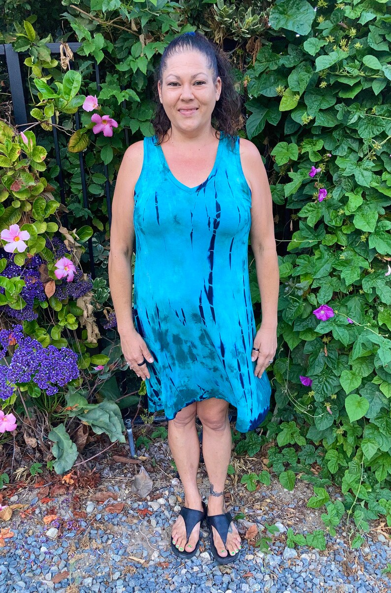 May include: A woman wearing a blue and green tie-dye sleeveless dress with a V-neck. She is standing in front of a green leafy background.