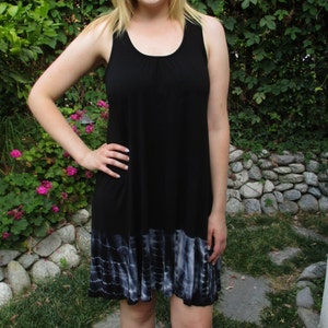 May include: A woman wearing a black sleeveless dress with a tie-dye hem. The dress has a scoop neckline and a relaxed fit.