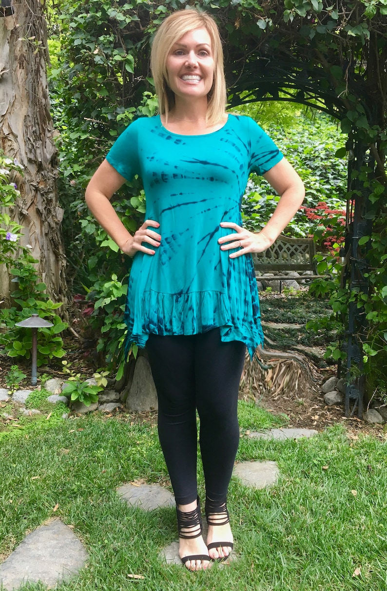 May include: A woman wearing a teal blue tie-dye shirt with a ruffled hem and black leggings. She is standing on a path with green grass and stones.