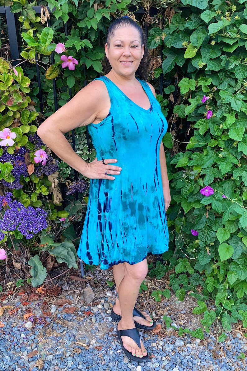 May include: A woman wearing a sleeveless blue tie-dye dress with a V-neckline. She is standing in front of a green leafy background.