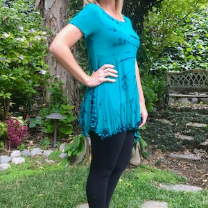 May include: A woman wearing a teal blue tie-dye shirt with a ruffled hem and black leggings. She is standing in a garden setting with green grass and a stone path.