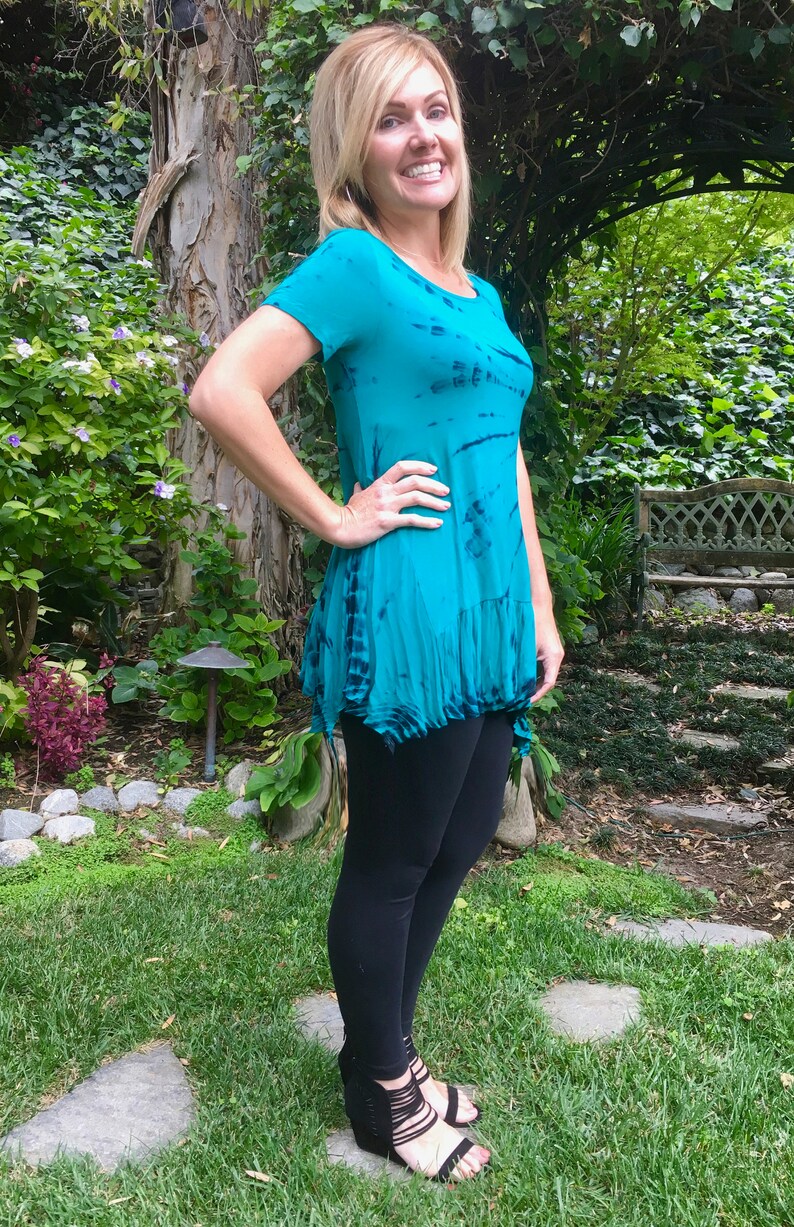 May include: A woman wearing a teal blue tie-dye shirt with a ruffled hem and black leggings. She is standing in a garden setting with green grass and a stone path.