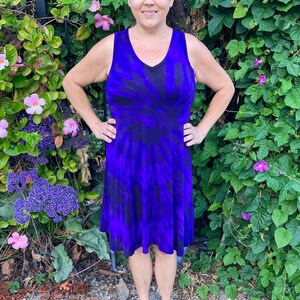 May include: A woman wearing a purple and black tie-dye sleeveless dress with a v-neck. She is standing in front of a green leafy background with pink flowers.