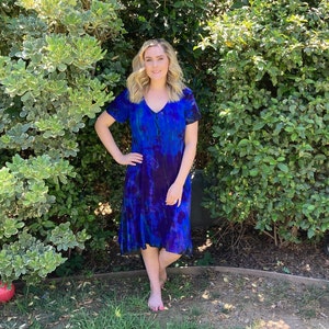 May include: A woman wearing a blue and purple tie-dye dress with a V-neck and short sleeves. She is standing in front of a green bush.