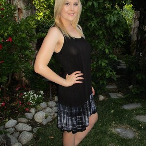 May include: A woman wearing a black sleeveless dress with a white tie-dye hem. The dress is loose-fitting and has a casual style. The woman is standing in a garden setting with green grass and rocks.