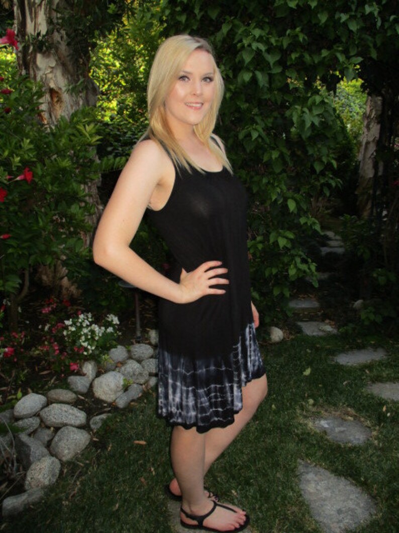 May include: A woman wearing a black sleeveless dress with a white tie-dye hem. The dress is loose-fitting and has a casual style. The woman is standing in a garden setting with green grass and rocks.