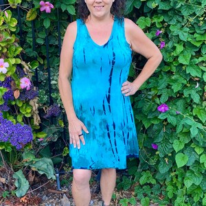May include: A woman wearing a sleeveless blue tie-dye dress with a V-neckline. She is standing in front of a green leafy background.
