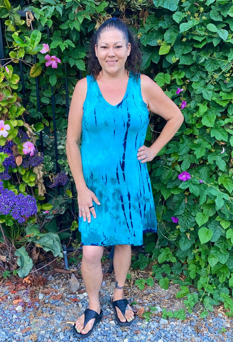 May include: A woman wearing a sleeveless blue tie-dye dress with a V-neckline. She is standing in front of a green leafy background.