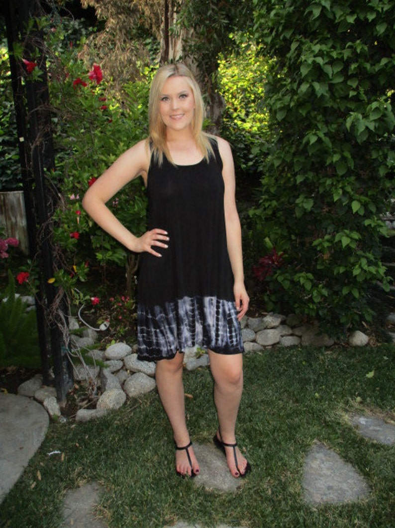 May include: A woman wearing a black sleeveless dress with a tie-dye hem. The dress has a scoop neck and a relaxed fit. She is standing in a garden setting.