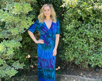 May include: A woman wearing a long, blue and purple tie-dye maxi dress with a V-neckline and short sleeves. The dress has a swirling pattern and is made of a soft, flowing fabric.