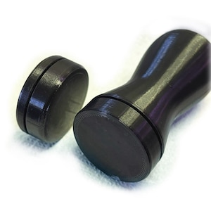 May include: A black coffee tamper with a smooth, contoured handle and a flat, circular base, accompanied by a separate, smaller black circular disc component. Both items feature a visible 3D printed texture with fine, concentric circular lines.