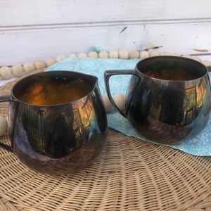 May include: Two antique silver-plated serving pieces. One is a creamer with a spout, and the other is a sugar bowl with two handles. Both have a dark, reflective finish with a gold interior. They sit on a woven wicker mat.
