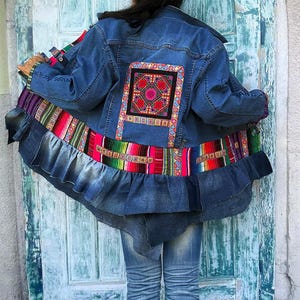 Könnte beinhalten: Eine blaue Jeansjacke mit einem großen, quadratischen, mehrfarbigen Stickaufnäher auf der Rückseite. Die Jacke hat einen breiten, gerüschten Jeanssaum und ein horizontales Band aus bunt gestreiftem Stoff am unteren Rücken.