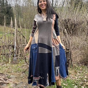 Op de afbeelding: Een patchwork jurk in tinten grijs, blauw en bruin. De jurk heeft een verscheidenheid aan stoffen texturen en denim zakken. De jurk met lange mouwen wordt gedragen met olijfgroene laarzen. De achtergrond omvat een houten hek en bomen.