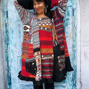 Op de afbeelding: Een tuniek met lange mouwen voorzien van een patchworkontwerp met diverse geometrische en gestreepte patronen in rood, zwart, wit en oranje. Het kledingstuk bevat twee voorzakken, een geplooide zoom en effen zwarte stoffen panelen op de mouwen.